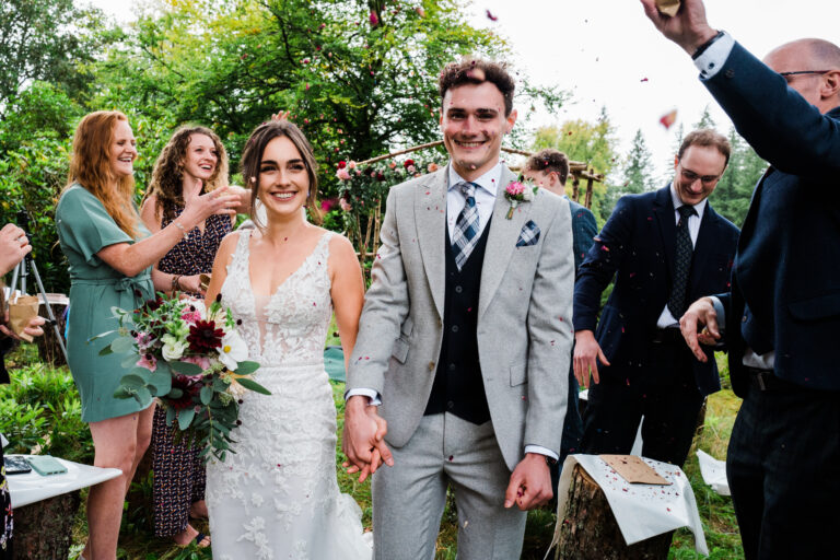 Bride and groom smile as the walk down a woodland aisle with confetti been thrown